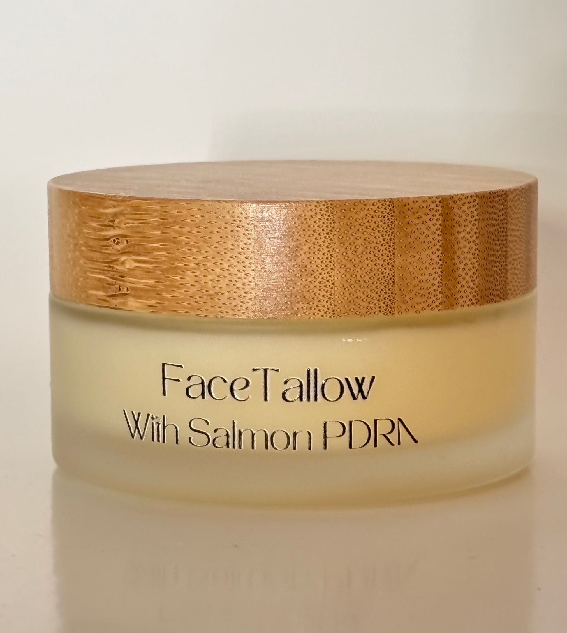 A jar of FaceTallow skincare cream with salmon PDRN, featuring a frosted glass container with pale yellow cream inside and a textured light brown bamboo lid, set against a plain off-white background.